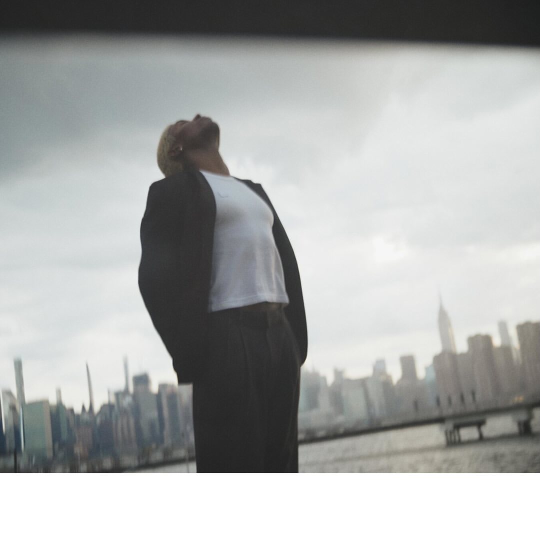 Adult with blond hair in a black blazer and white shirt looking up at the gray, cloudy sky with a city skyline in the background.