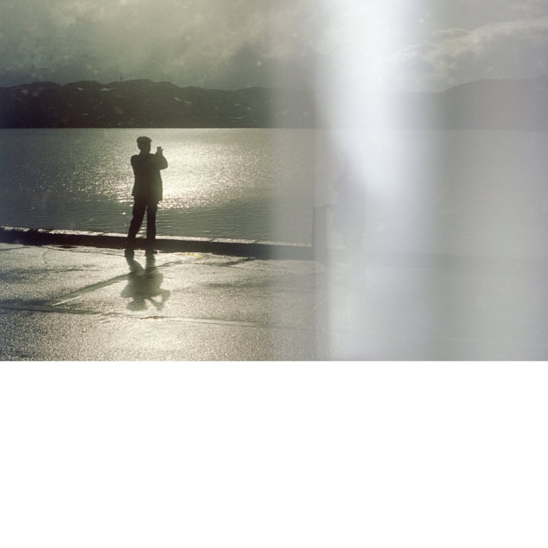Man stands silhouetted by a reflective body of water against a hazy mountain backdrop, creating a contemplative and scenic view.