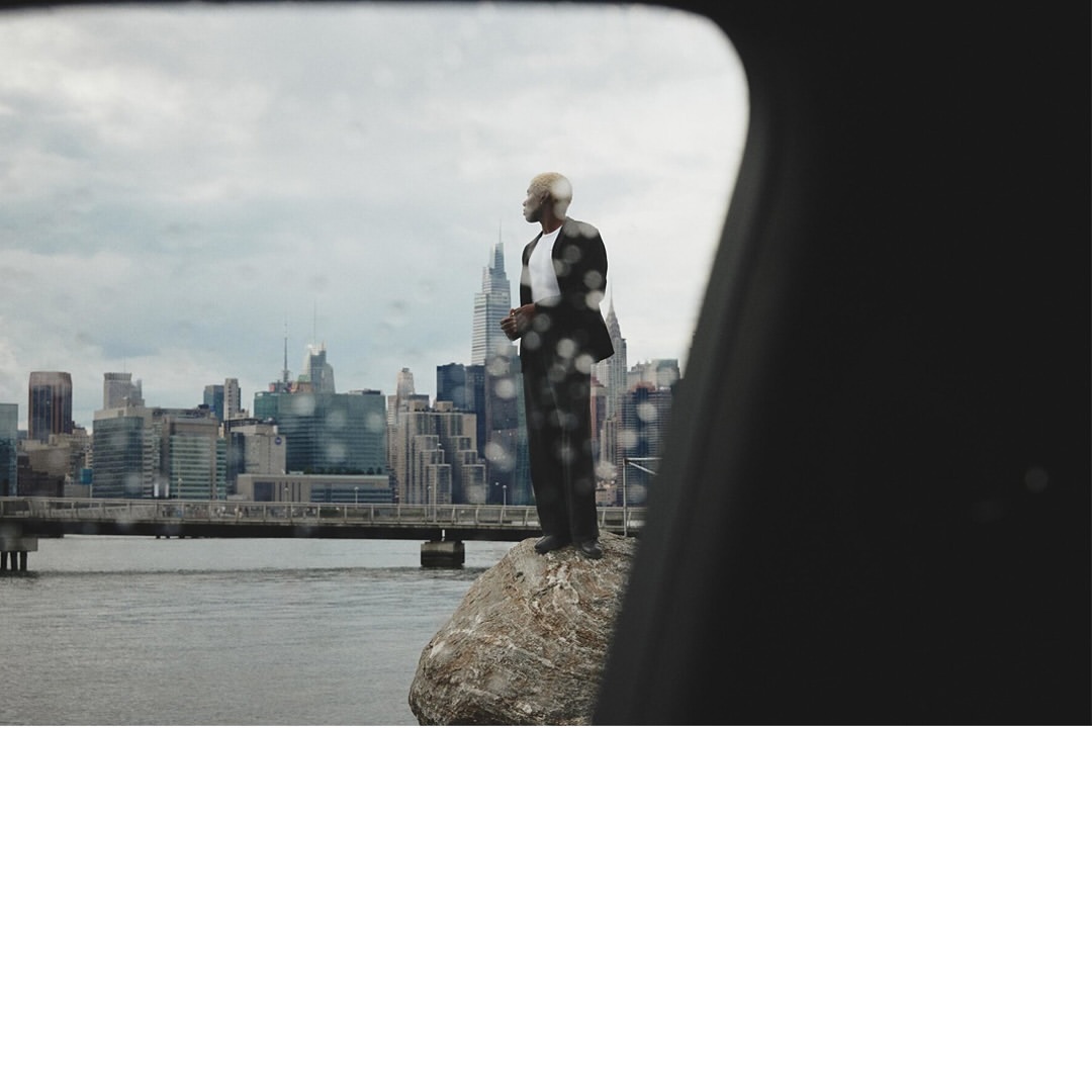 A man in a suit standing on a rock with city skyline in the background, viewed through a frame with raindrops on glass.