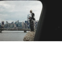 A man in a black suit standing on a rock by a river with a city skyline in the background, viewed through a rain-spotted window frame.