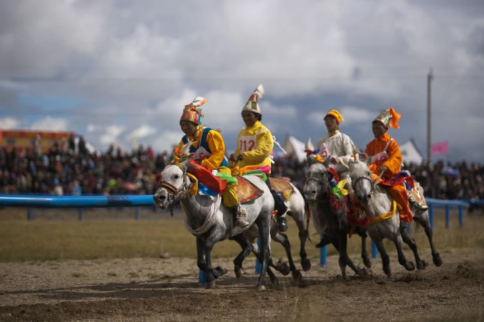 Four horse riders in bright tunics, racing across a dirt field in front of a large crowd, under a partly cloudy sky.
