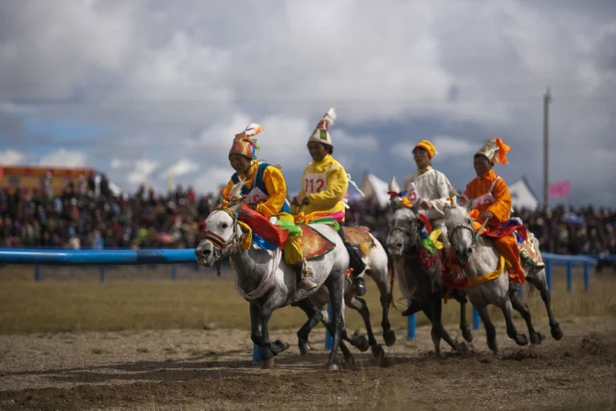 Four horse riders in bright tunics, racing across a dirt field in front of a large crowd, under a partly cloudy sky.