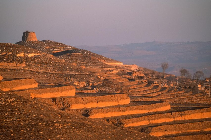 Terraced farmland on hillsides with a small stone structure on the hilltop under a clear sky, creating a rural landscape scene.