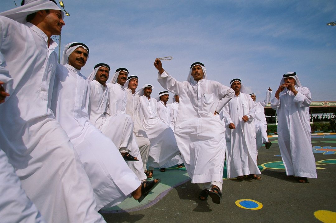Group of adult males in traditional white clothing dancing outdoors under a sunny sky, with a man holding beads in his raised hand.