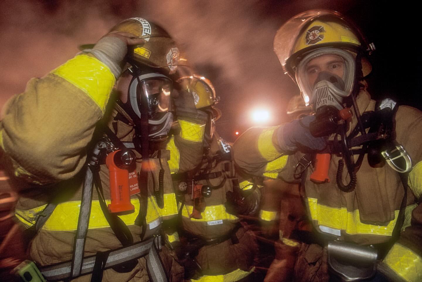 Three firefighters in protective gear and helmets with face masks stand in a smoky environment with bright yellow reflective stripes.