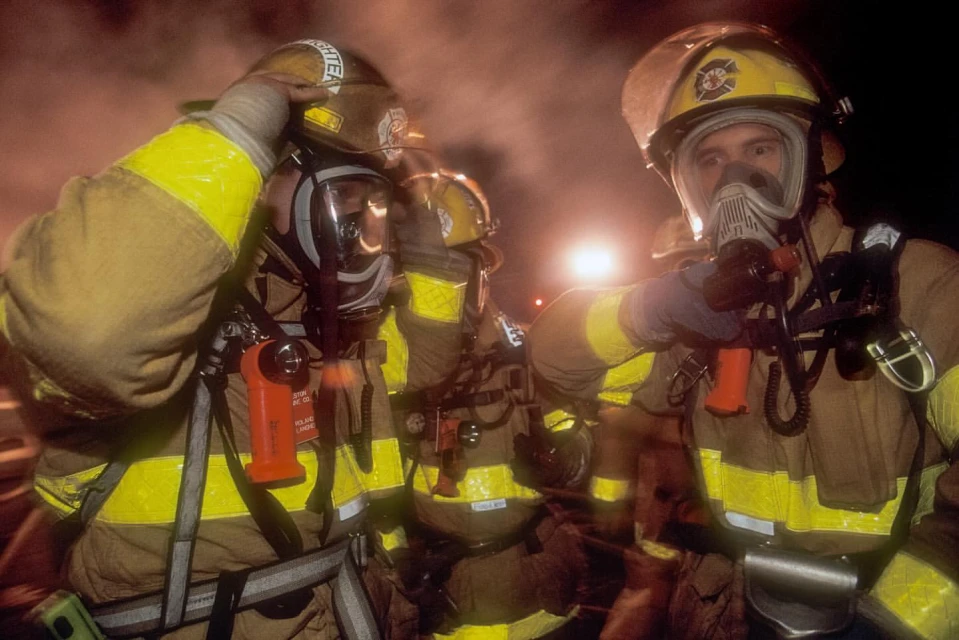 Three firefighters in protective gear and helmets with face masks stand in a smoky environment with bright yellow reflective stripes.