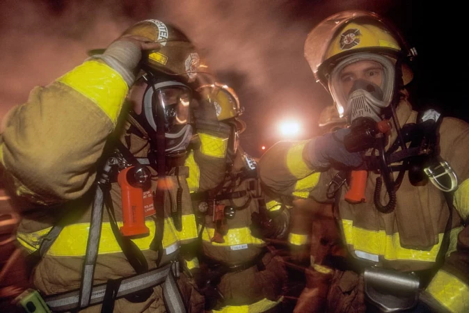 Three firefighters in protective gear and helmets with face masks stand in a smoky environment with bright yellow reflective stripes.