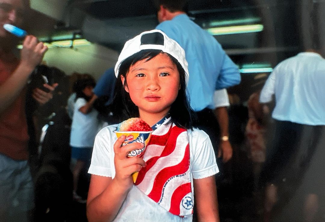 A young girl in a white hat holds a cup of shaved ice, wearing an American flag scarf in an indoor setting with a crowd in the background.