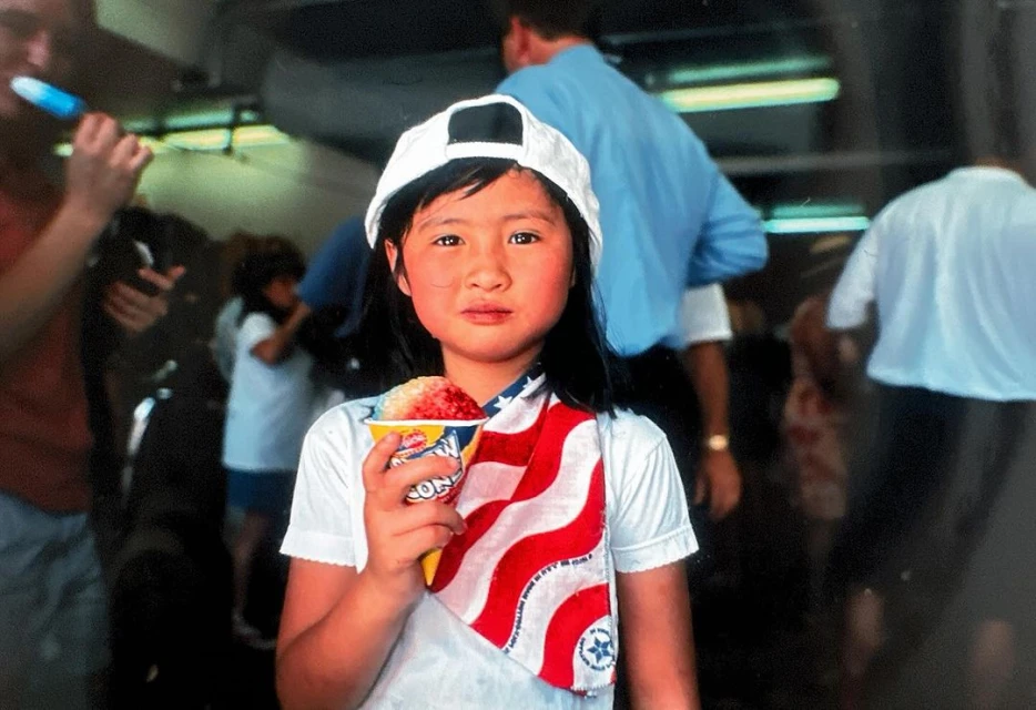 A young girl in a white hat holds a cup of shaved ice, wearing an American flag scarf in an indoor setting with a crowd in the background.