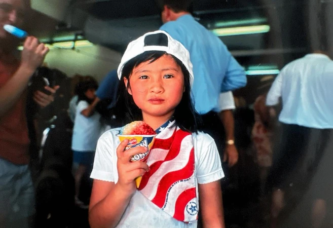 A young girl in a white hat holds a cup of shaved ice, wearing an American flag scarf in an indoor setting with a crowd in the background.