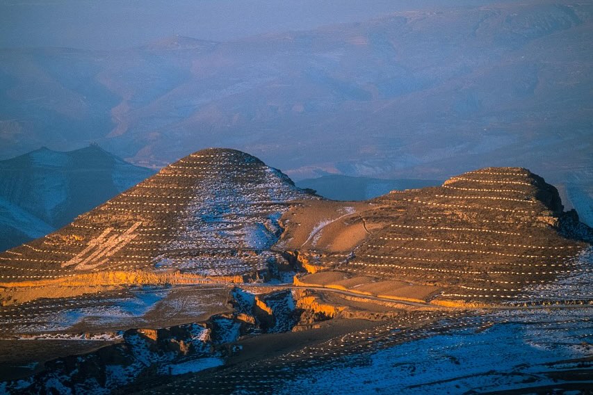 Scenic landscape featuring terraced mountains with a dusting of snow, under a soft, golden-blue light, conveying a serene atmosphere.