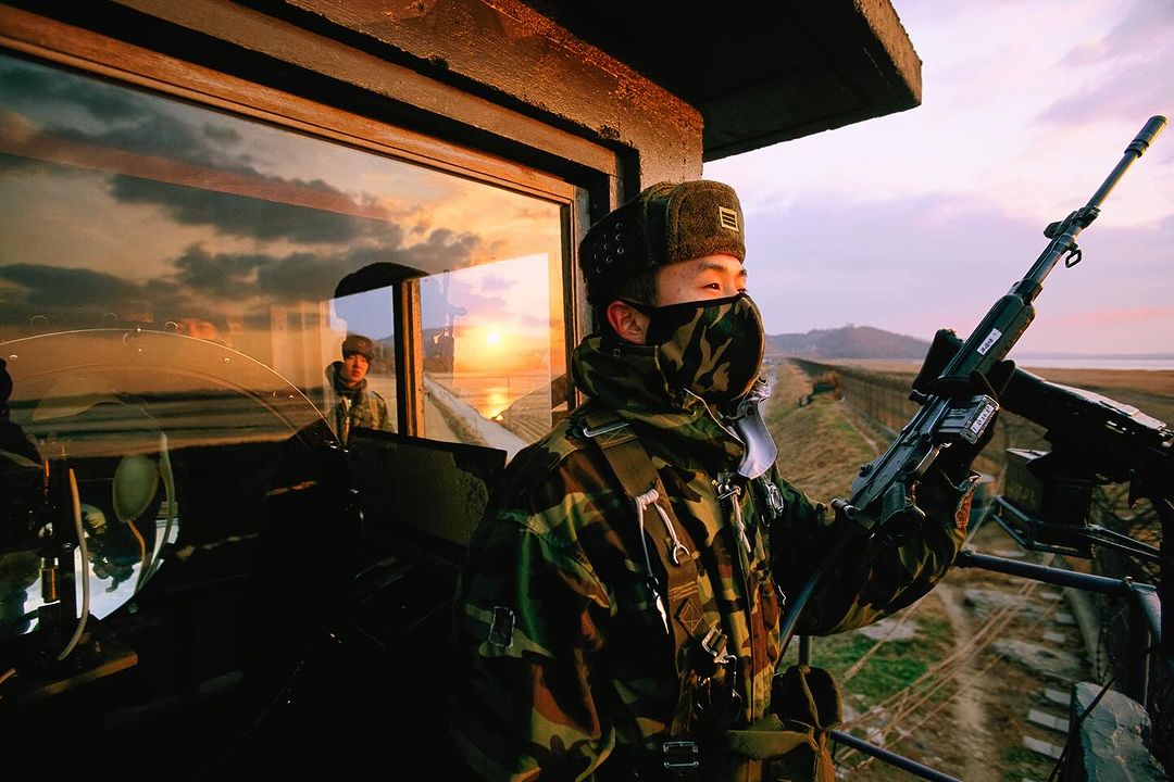 Soldier in camouflage uniform holding rifle in a watchtower at sunset, looking out over a fence and distant landscape in a patrol.