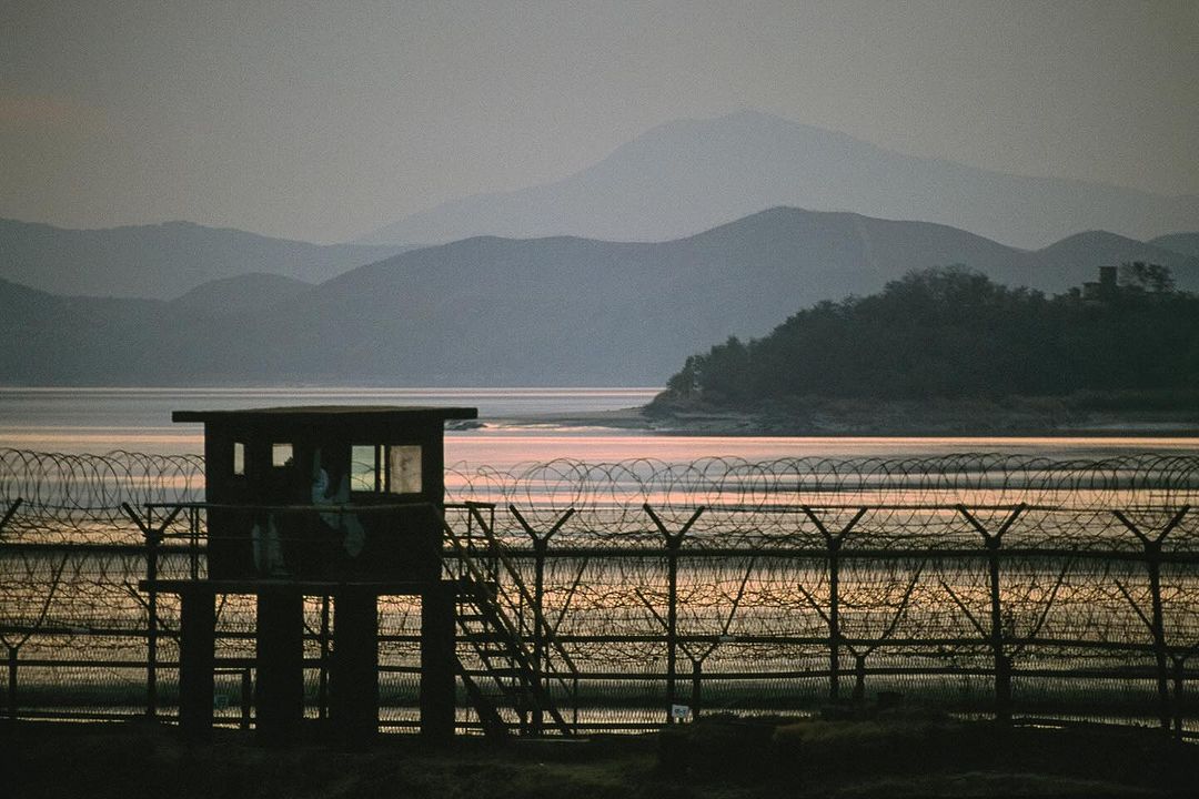 Barbed wire fence and watchtower on the water against mountains in the background under a dusky sky at dawn or dusk.