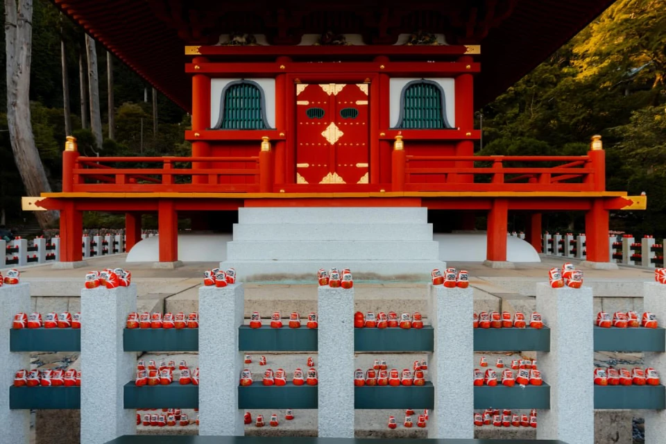 A three-tiered platform with red Japanese architecture and rows of red Daruma dolls at the base, surrounded by greenery and trees.
