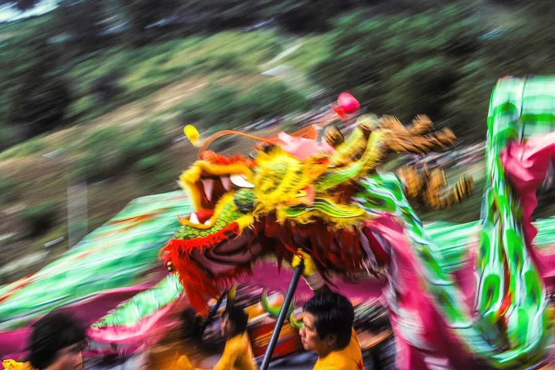 Chinese Dragon Dance parade featuring vibrant colors and blurred motion, set against a backdrop of green hills and a sunny day.