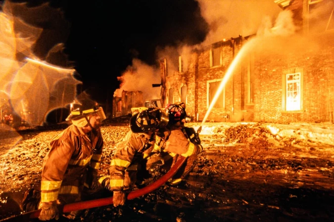 Three firefighters in protective gear battle a fire at night with a water hose. Smoke billows around a brick building.