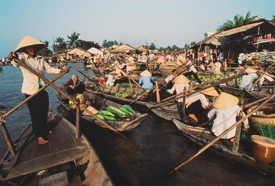 A bustling river market in Southeast Asia featuring many long wooden boats filled with vendors and customers, houses lining the riverbank.