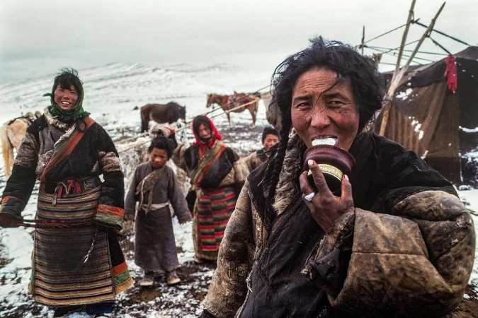 Group of people in traditional clothing standing on a snowy field, with horses and tents in the background, drinking and smiling.