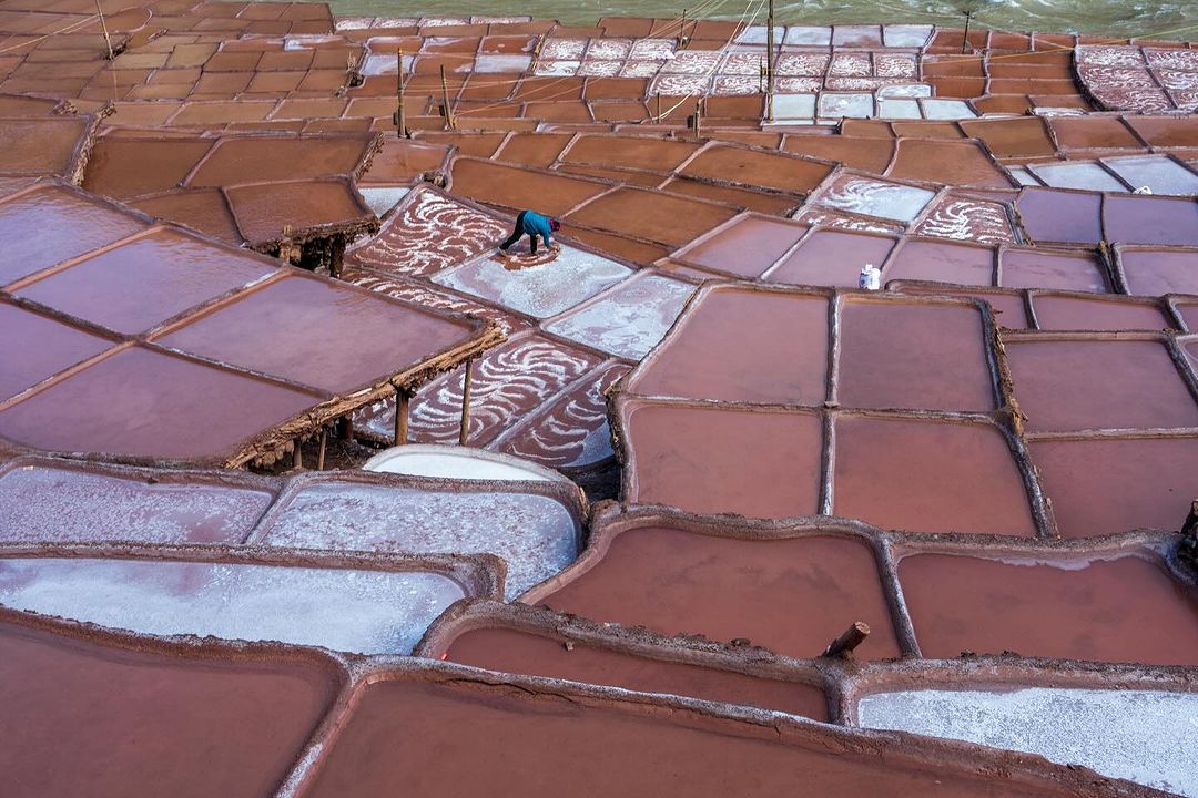 Aerial view of the Maras Salt Mines in Peru shows a person working, with layered salt pans filled with pink and white water.
