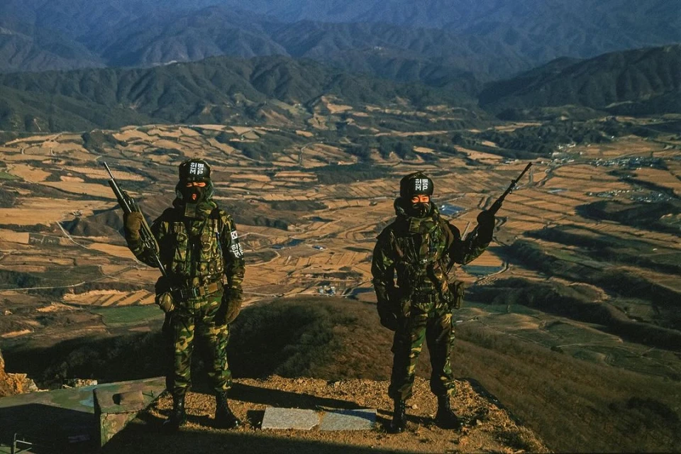 Two masked soldiers in camouflage uniforms stand guard on a mountain peak overlooking a valley, holding rifles, with mountains in the background.