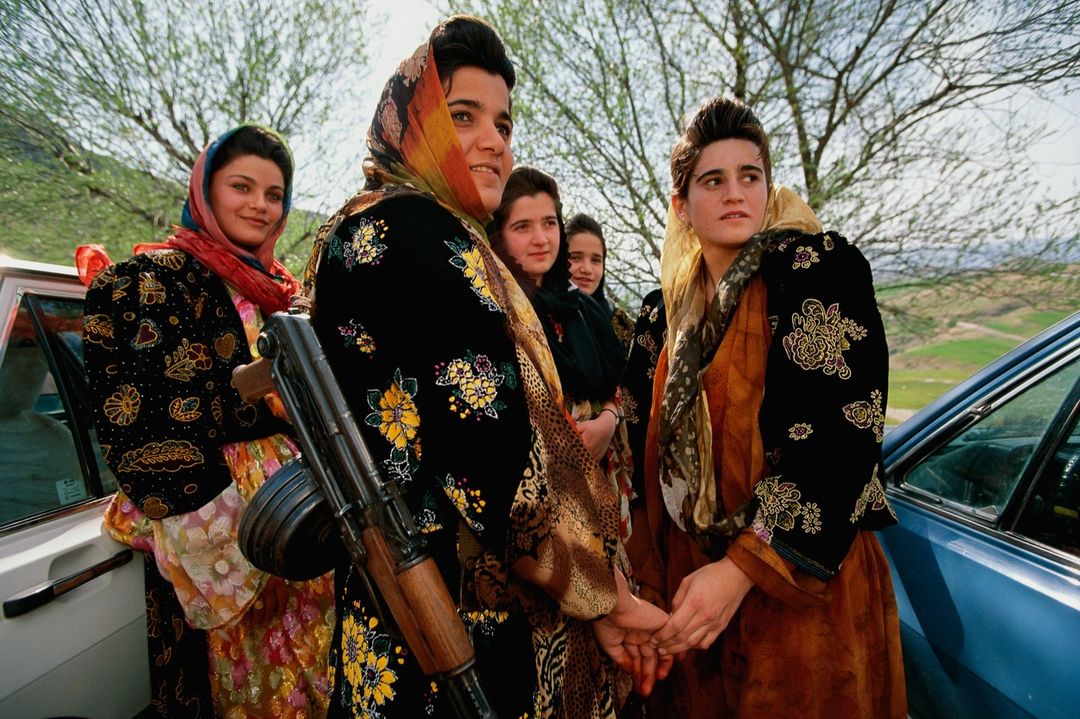Group of women in traditional dress and headscarves standing outside near a car and a green, hilly landscape with trees.