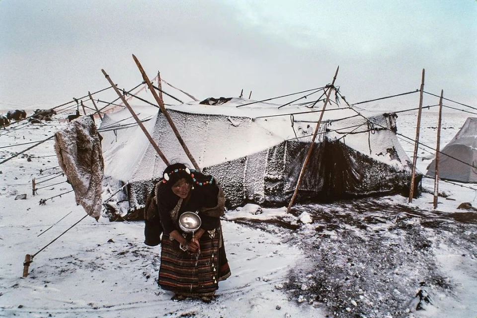 Adult woman in traditional dress stands outside snow-covered tents in a rural, cloudy environment, holding a metal bowl.