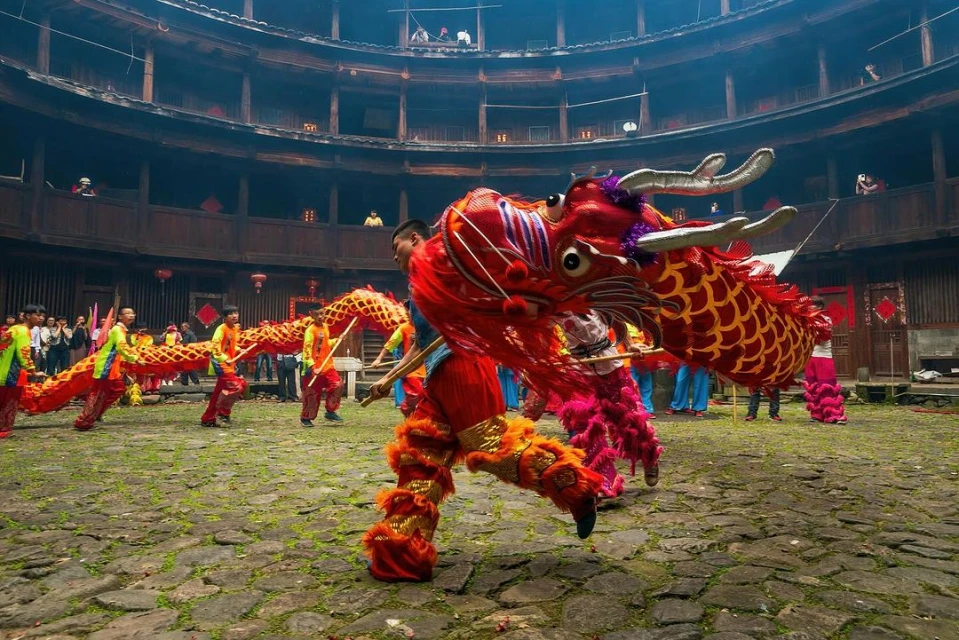 People perform a traditional dragon dance in a round, multi-story building courtyard, featuring red and gold dragon costumes and a stone floor.
