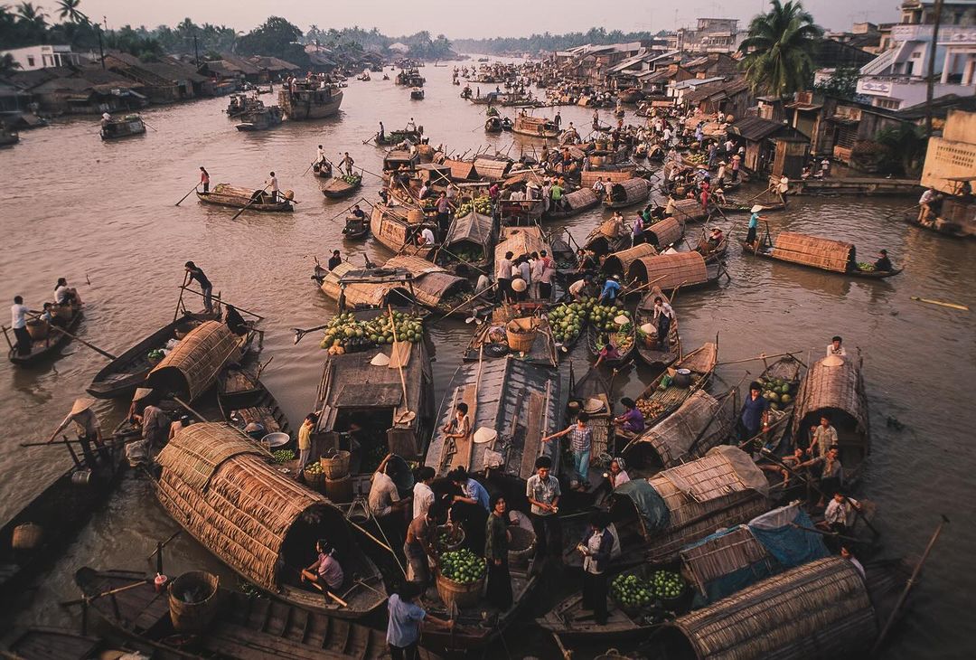 Busy floating market in Southeast Asia featuring many wooden boats filled with produce, adults trading, and houses along the riverbank.