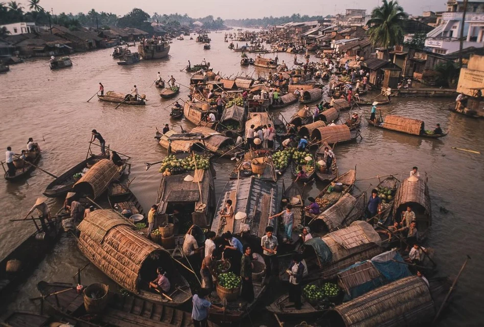 Busy floating market in Southeast Asia featuring many wooden boats filled with produce, adults trading, and houses along the riverbank.