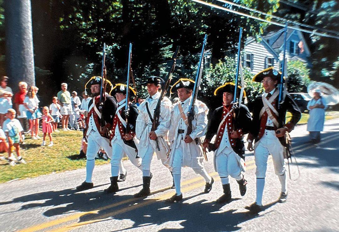Six men in Revolutionary War uniforms marching in a parade on a road, with a crowd and houses visible in the background.