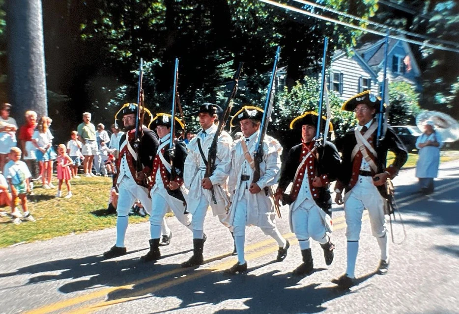 Six men in Revolutionary War uniforms marching in a parade on a road, with a crowd and houses visible in the background.