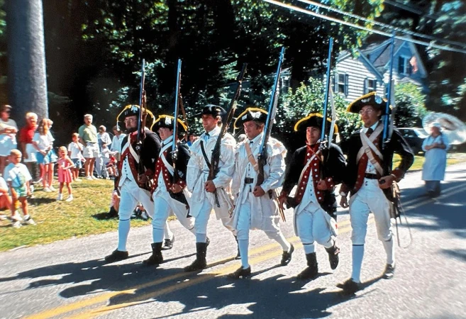 Six men in Revolutionary War uniforms marching in a parade on a road, with a crowd and houses visible in the background.