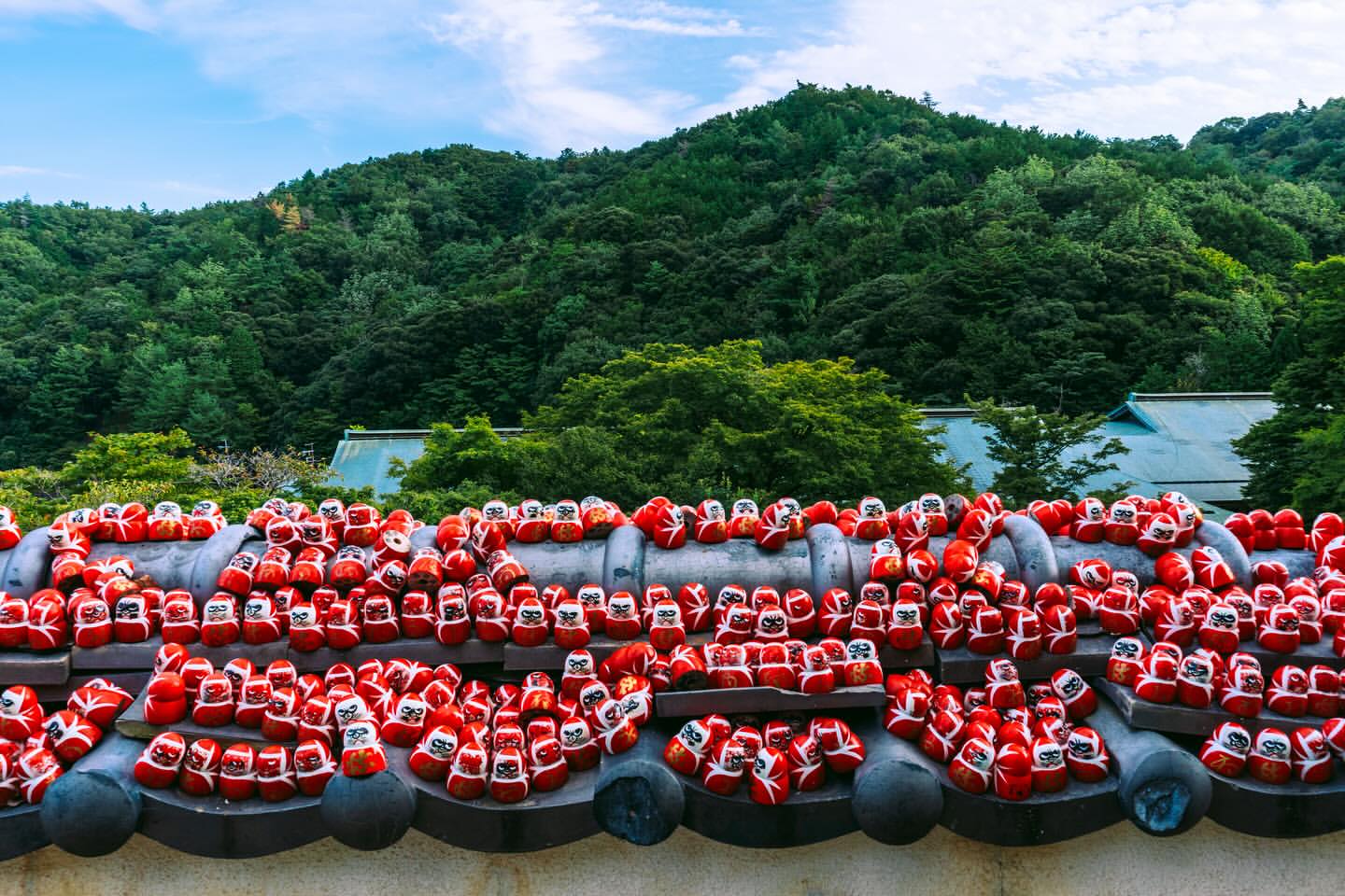 Rows of red Daruma dolls line a stone wall in front of a lush green mountainside, with a blue sky above in an outdoor setting.
