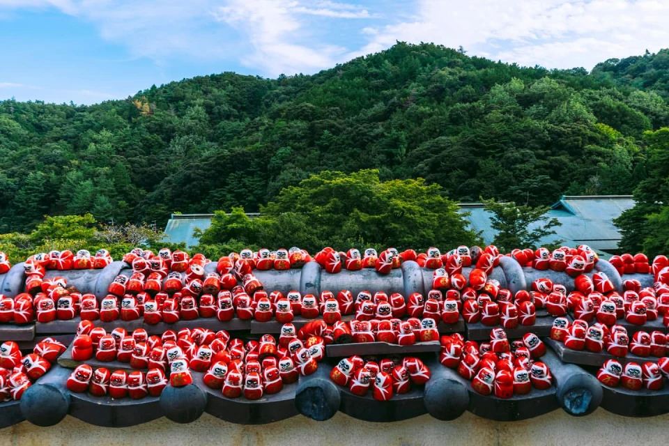 Rows of red Daruma dolls line a stone wall in front of a lush green mountainside, with a blue sky above in an outdoor setting.