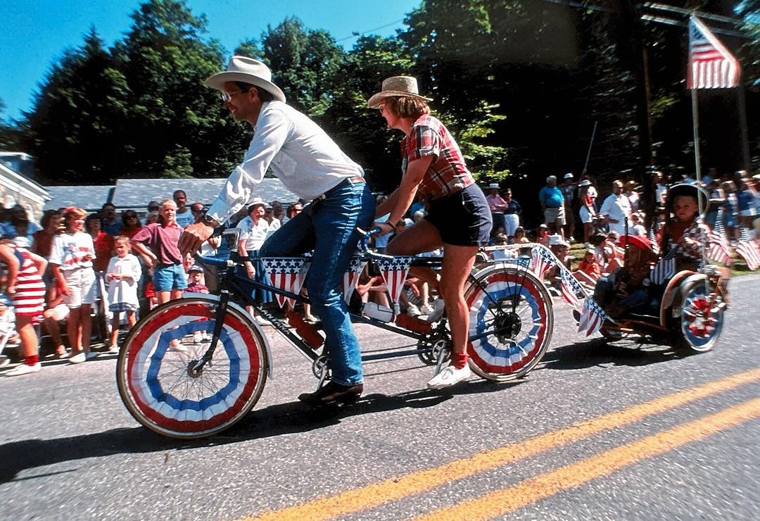 Tandem bicycle decorated with American flags and bunting being ridden by two adults and pulling a child in a small trailer in a parade.
