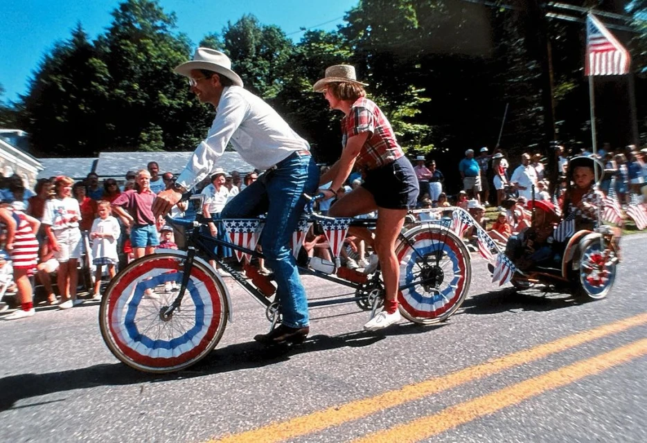 Tandem bicycle decorated with American flags and bunting being ridden by two adults and pulling a child in a small trailer in a parade.