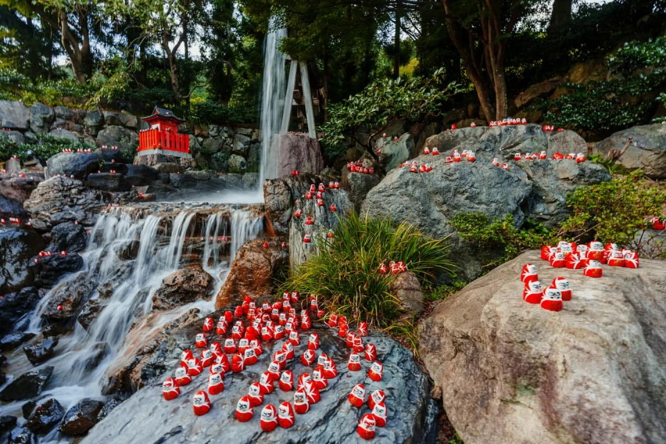Scenic view of a waterfall cascading over rocks adorned with miniature red Daruma dolls, set against a backdrop of lush greenery.