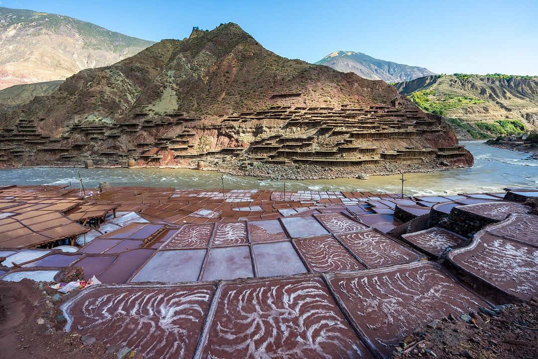 Terraced salt ponds and brown earth buildings are set against a mountain and river landscape under a bright blue sky in this rural outdoor scene.