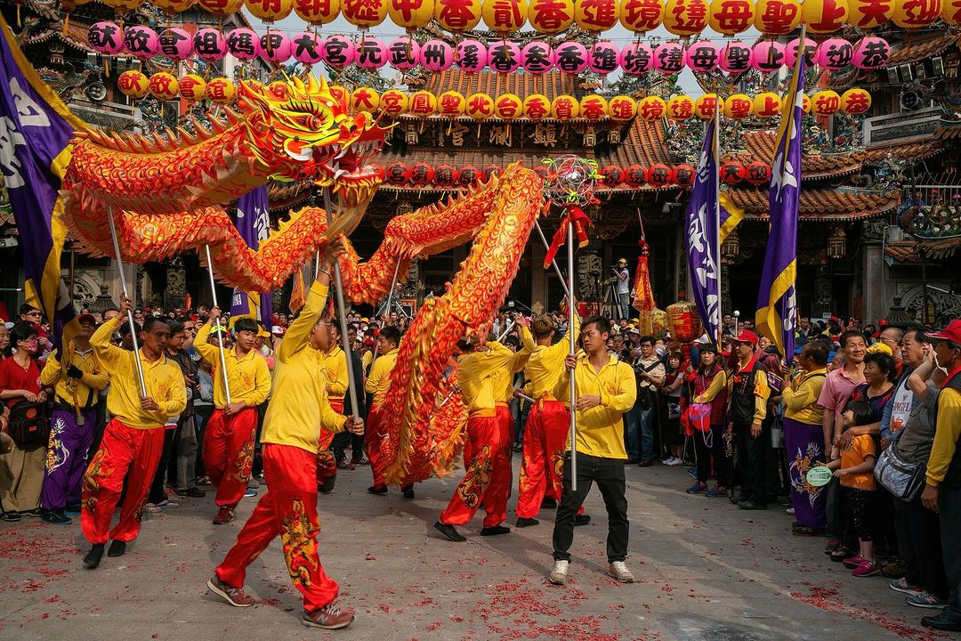 A dragon dance performance with a large crowd in front of a temple adorned with lanterns, showcasing a traditional cultural celebration.