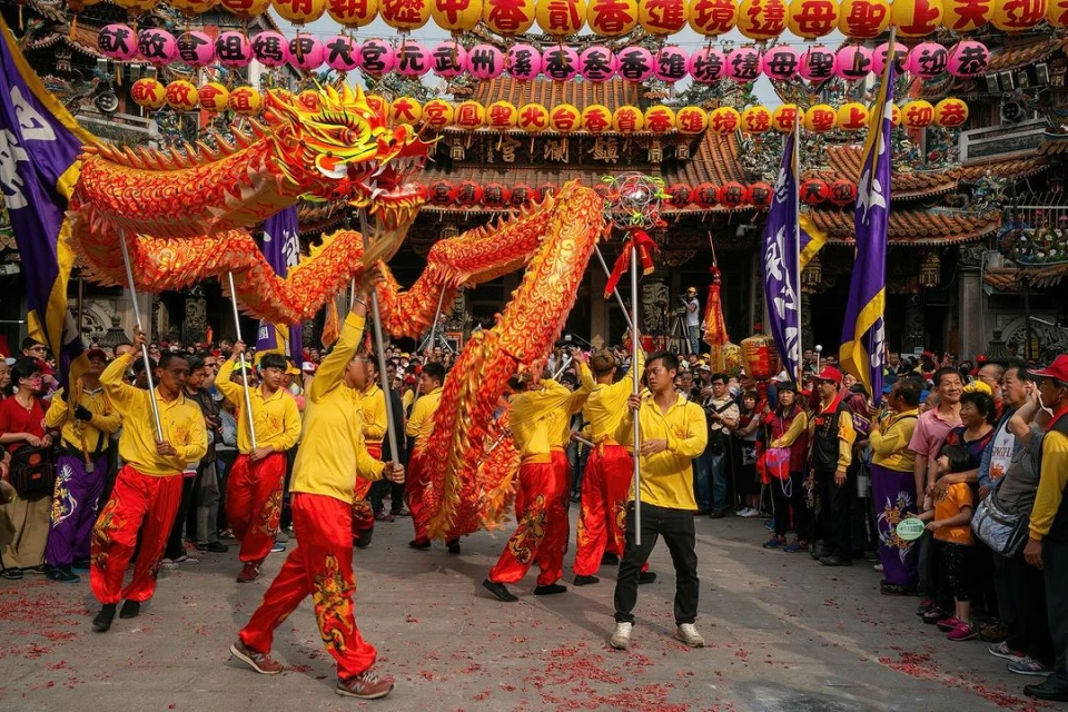 A dragon dance performance with a large crowd in front of a temple adorned with lanterns, showcasing a traditional cultural celebration.