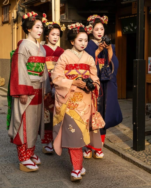 Four Japanese women in kimonos stand together outdoors. Three hold cameras; they are wearing traditional makeup, ornate hairstyles, and wooden shoes.