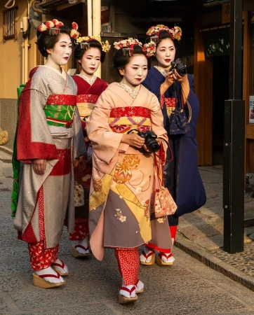 Four Japanese women in kimonos stand together outdoors. Three hold cameras; they are wearing traditional makeup, ornate hairstyles, and wooden shoes.