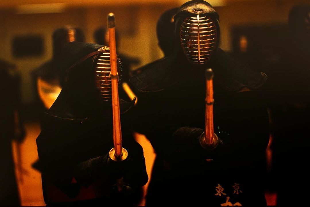 Two kendo fighters wearing protective helmets and dark clothing, holding swords in a dimly lit setting, focus on martial arts combat.
