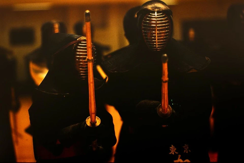 Two kendo fighters wearing protective helmets and dark clothing, holding swords in a dimly lit setting, focus on martial arts combat.