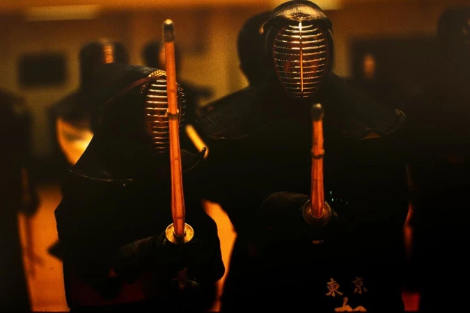 Two kendo fighters wearing protective helmets and dark clothing, holding swords in a dimly lit setting, focus on martial arts combat.