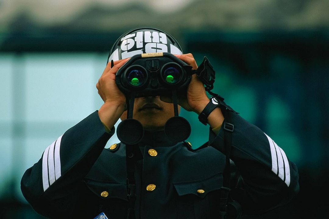 An individual in a dark uniform and helmet gazes through binoculars. The person is the primary focus, set against a blurred backdrop.
