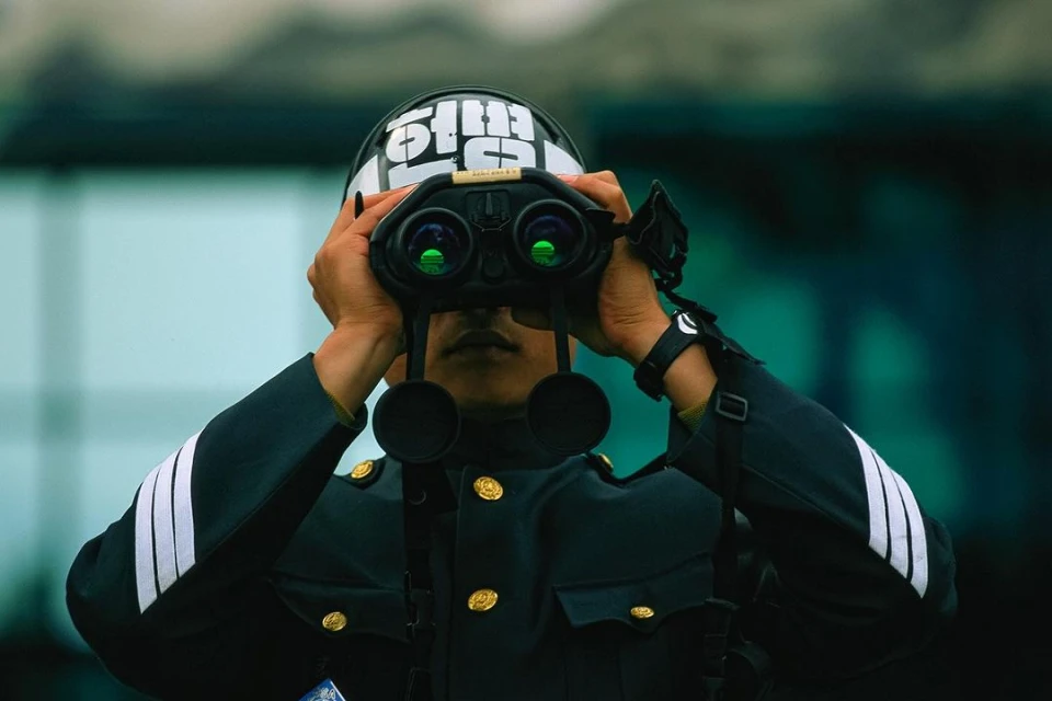 An individual in a dark uniform and helmet gazes through binoculars. The person is the primary focus, set against a blurred backdrop.