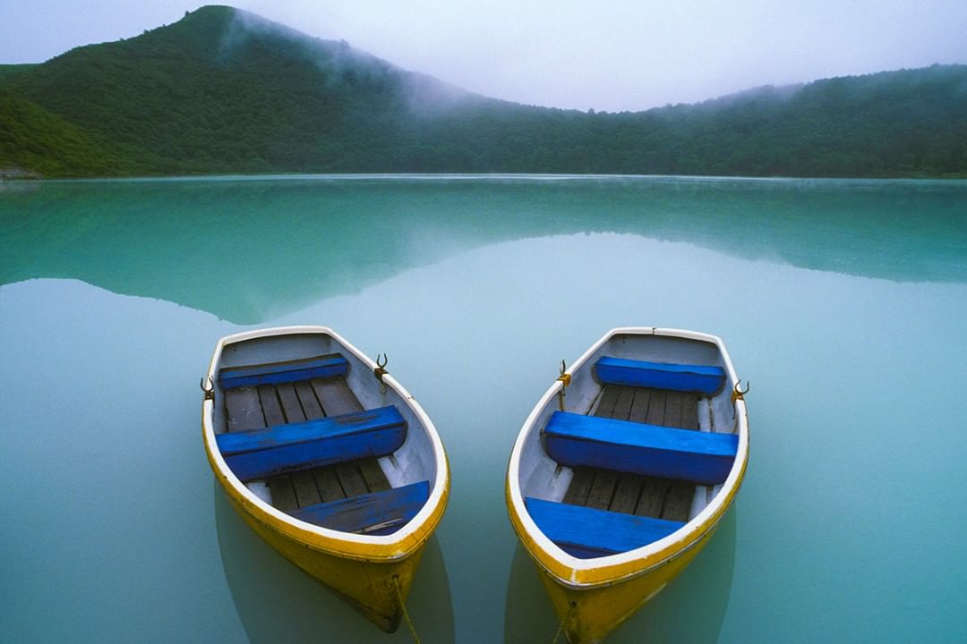Two yellow and white rowboats anchored in a tranquil turquoise lake, with a backdrop of a green mountain covered in mist.
