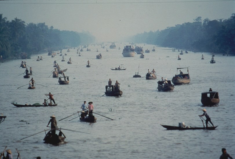 Numerous traditional boats navigate a wide river in Asia, with lush green trees lining the banks under a soft, overcast sky.