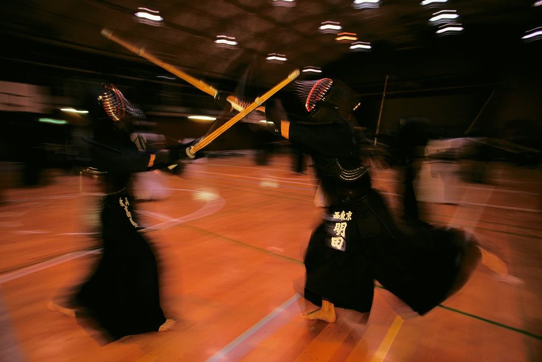 Two kendo fighters in protective gear compete in a dimly lit indoor space with blurred motion, wielding bamboo swords in a clash.